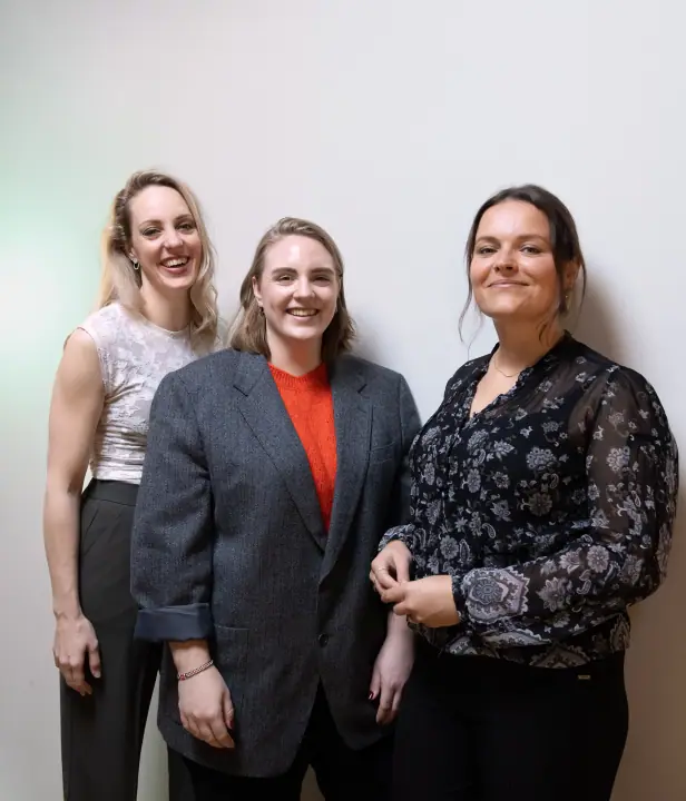 Groepsportret van drie vrouwen naast elkaar tegen een lichte muur, alle drie lachend
