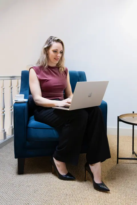 Vrouw werkt op haar laptop in een blauwe fauteuil, met hakken aan en een kopje koffie naast zich