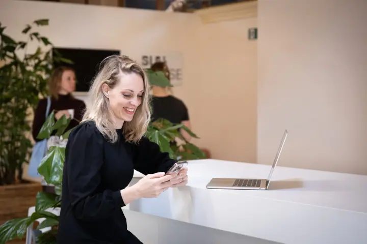 Vrouw kijkt glimlachend op haar telefoon aan een witte balie, met twee mensen tussen de planten erachter