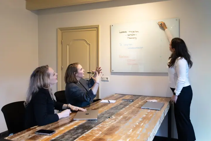 Vrouw schrijft op een glazen whiteboard terwijl twee anderen vanaf de tafel meedenken