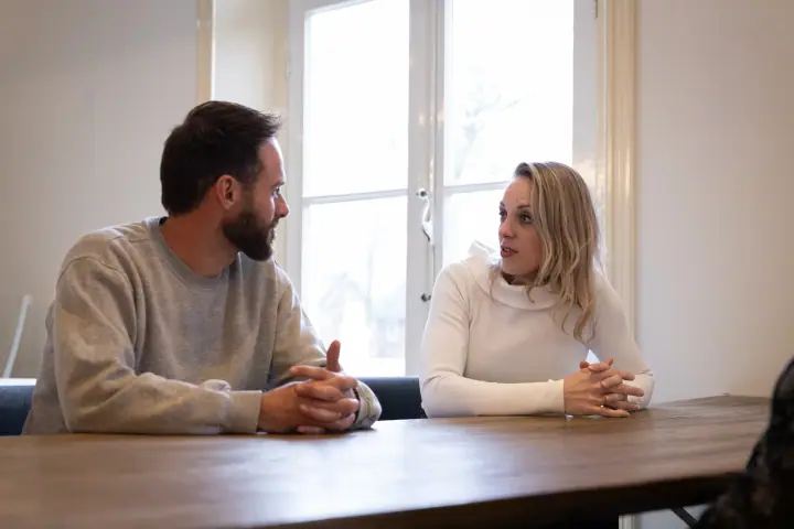 Man met baard en blonde vrouw in witte coltrui praten met gevouwen handen aan tafel
