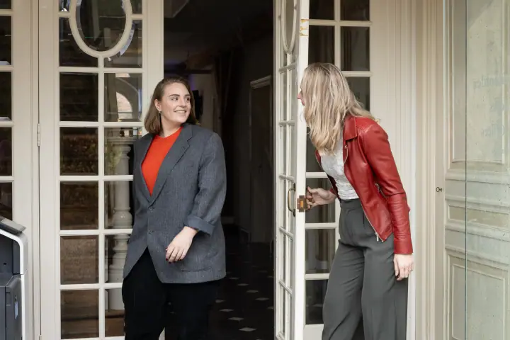 Vrouw in grijs colbert ontvangt een bezoeker in rood leren jasje bij de glazen voordeur