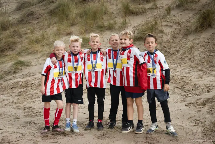 Zes jongens in rood-wit gestreepte voetbalshirts met medaille en startnummer, arm in arm voor een duin