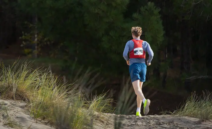 Trailrunner met rood hesje en startnummer loopt een zandpad in de duinen op, richting de dennen