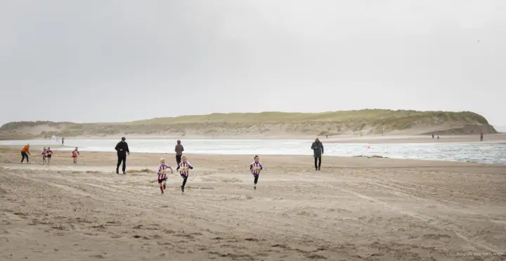 Kinderen in gestreepte shirts rennen over het brede strand bij Mudsweattrail 2025, ouders lopen mee