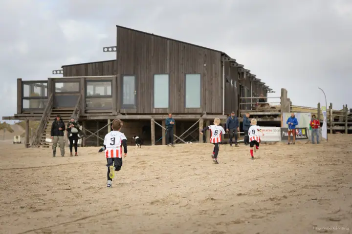 Drie jonge lopers met rugnummer 3, 6 en 8 sprinten over het zand naar een houten strandpaviljoen