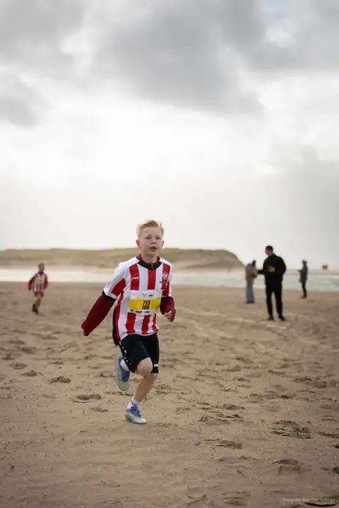 Jongen in rood-wit shirt met startnummer rent over het strand op de camera af, onder een grijze lucht