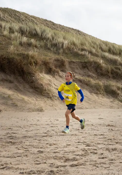 Meisje in geel shirt met startnummer loopt soepel over het zand onder een begroeide duinhelling