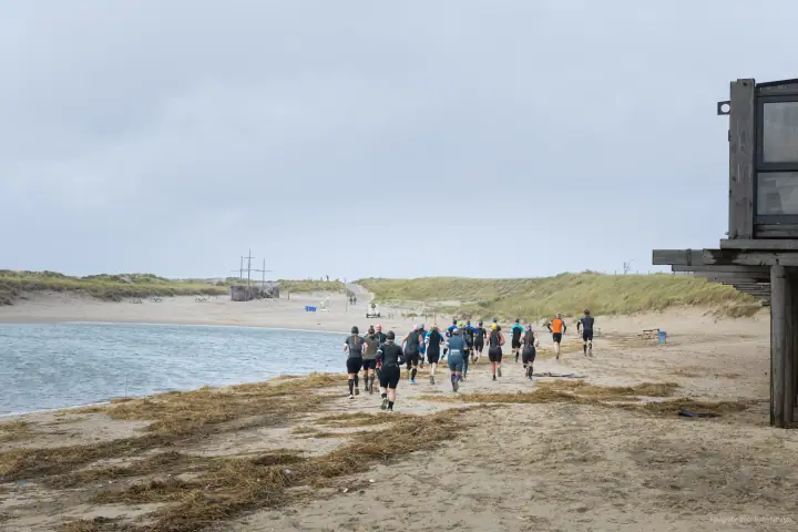 Een groep lopers in wetsuits trekt langs de waterlijn het strand af, richting de duinen