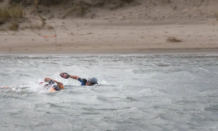 Twee zwemmers in wetsuits ploeteren door woelig water, een van hen met handpeddels