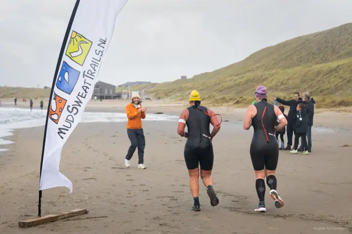 Twee deelnemers in wetsuit rennen na het zwemdeel over het strand langs de vlag van Mudsweattrails