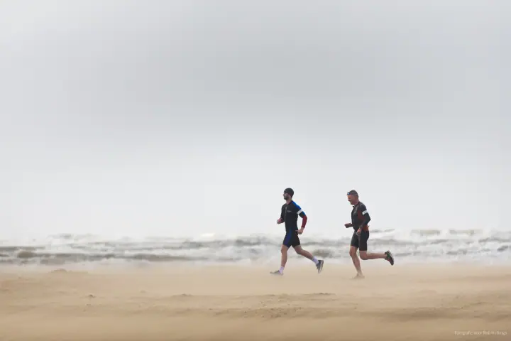Twee lopers in triatlonpakken rennen over het strand terwijl het zand om hun benen stuift