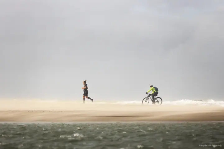 Een loper en een mountainbiker steken het strand over in een wolk opwaaiend zand