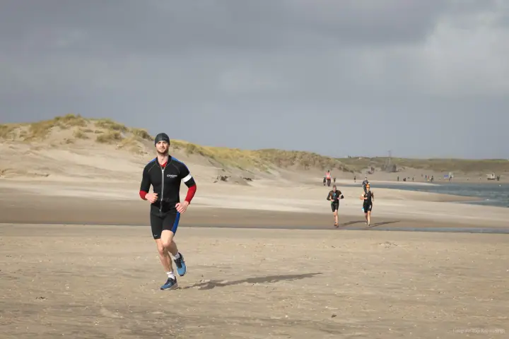 Loper in zwart pak met rode mouwen komt over het zandstrand aangelopen, duinen en donkere lucht erachter