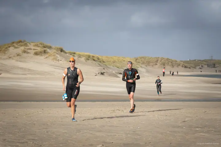 Twee deelnemers in wetsuits lopen over het strand naar de camera, een van hen met oranje badmuts