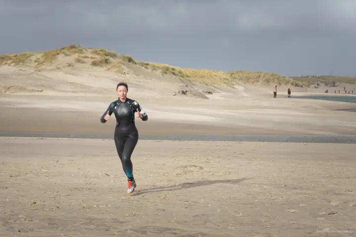 Een loopster in wetsuit steekt beide duimen op terwijl ze over het strand komt aanrennen