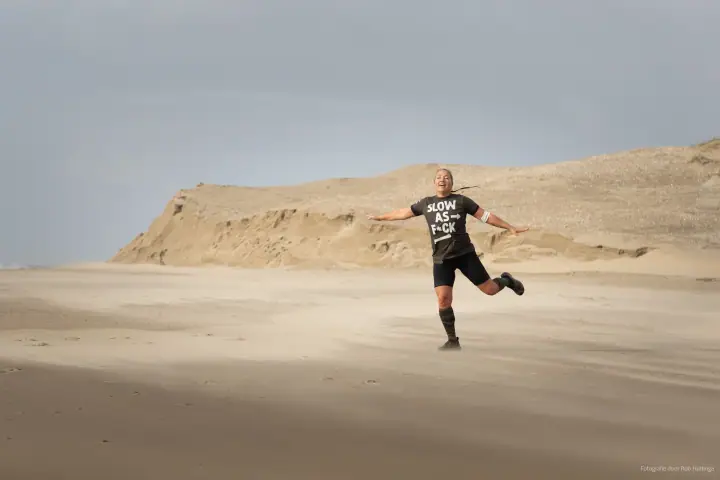 Een deelnemer springt uitgelaten op het strand met gespreide armen, een zandduin op de achtergrond