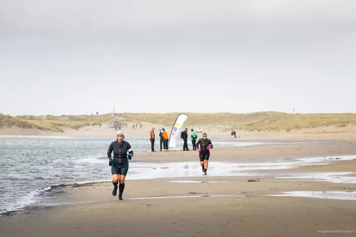 Twee lopers volgen de waterlijn, met toeschouwers en een beachvlag verderop