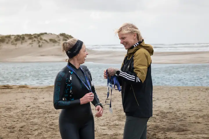Vrijwilliger hangt een medaille om bij een deelnemer in wetsuit, met de zee en de duinen erachter