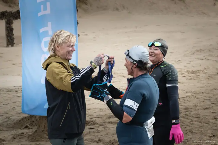 Een vrijwilliger hangt een medaille om bij een lachende finisher in badmuts, een tweede finisher lacht mee
