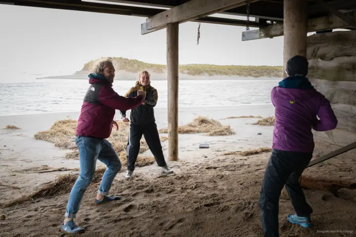 Onder het strandpaviljoen bewegen drie mensen uitbundig met hun armen, de zee op de achtergrond