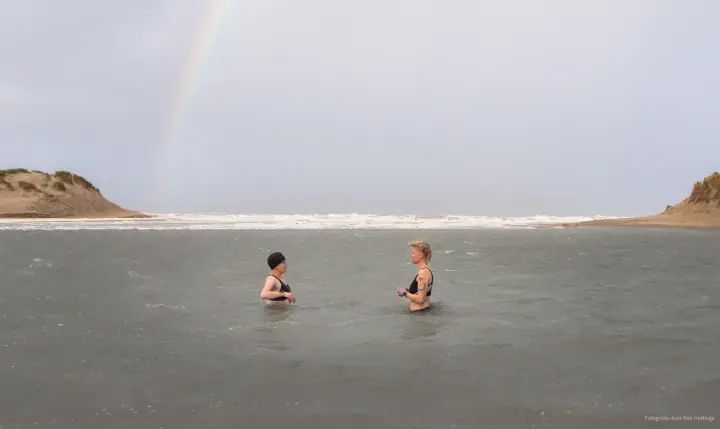 Twee vrouwen staan tot hun middel in het water, met een regenboog boven de duinen