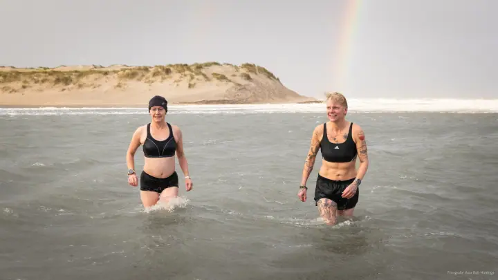 Twee deelnemers waden lachend uit zee bij Mudsweattrail 2025, met een regenboog boven het duin