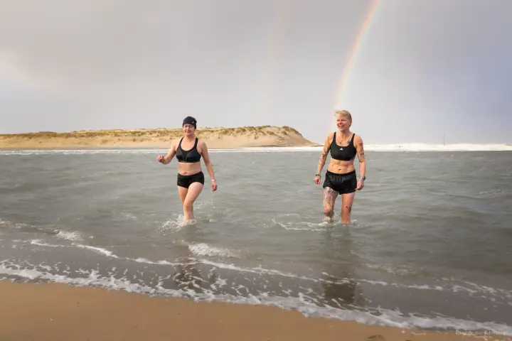 Twee vrouwen in sportkleding lopen lachend het water uit, een regenboog boven de duin