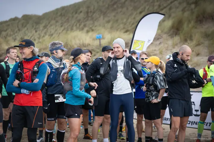 Trailrunners met hardloopvesten verzamelen zich bij de start, voor een duin vol helmgras