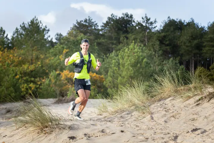 Trailrunner in fluogeel shirt steekt twee duimen op, op een zandpad tussen de dennen