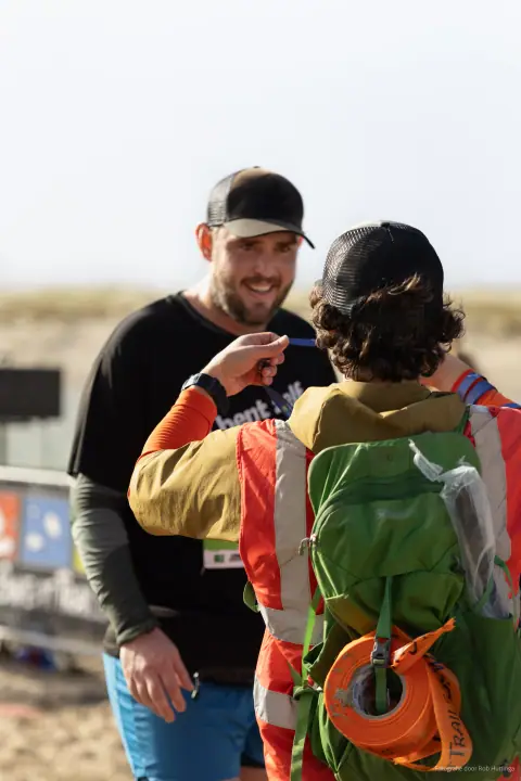 Een vrijwilliger in oranje hesje hangt een medaille om bij een lachende trailrunner met pet