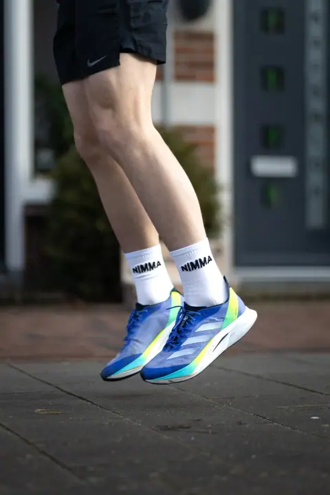 Beide voeten los van de grond, NIMMA sokken en blauwe schoenen scherp tegen een wazige gevel