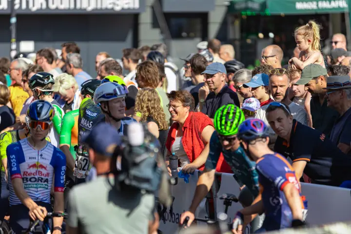Renners schuiven aan bij het dranghek voor de start, terwijl een cameraman filmt en het publiek dicht opeen toekijkt