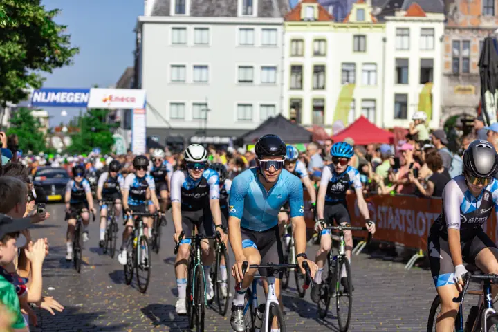 Renner in lichtblauw shirt rijdt voorop in een groep die publiek en startboog passeert