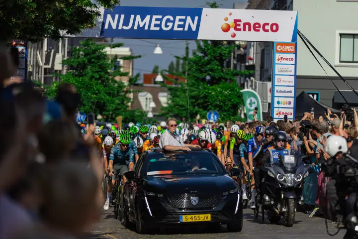 Auto met official in het dak leidt het peloton onder de startboog van het NK Wielrennen in Nijmegen
