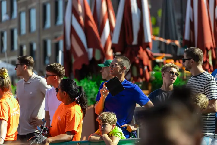 Toeschouwers en vrijwilligers in oranje shirts volgen de koers, een man filmt met zijn tablet