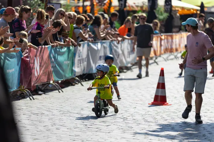 Peuter in geel Rabo-shirt rijdt op een loopfiets langs juichend publiek, een begeleider in roze shirt loopt mee