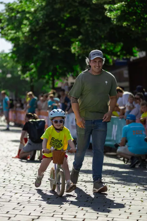 Kind met een mandje aan de loopfiets, met een lachende begeleider die ernaast meeloopt