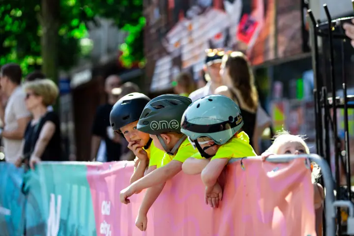 Kinderen in fluogele shirts leunen op een roze dranghek en kijken naar de race