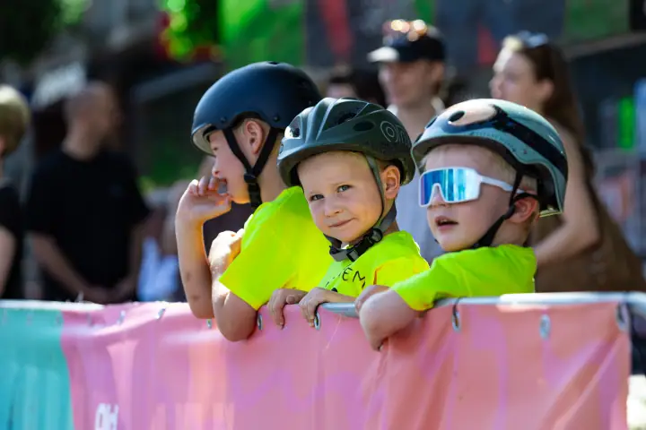Kind met groene helm kijkt lachend in de camera, naast twee andere kinderen aan het hek