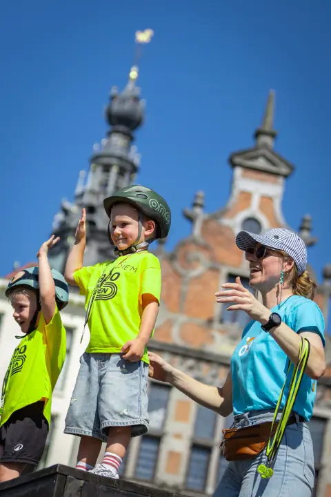 Twee kinderen zwaaien vanaf een podium, met een historische gevel tegen de blauwe lucht