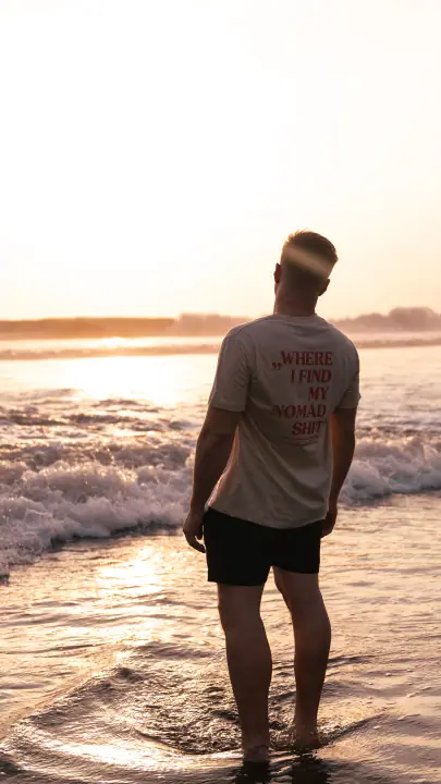 Tegen de ondergaande zon staat een man in de branding, de Nomadwise tekst rood op zijn beige shirt