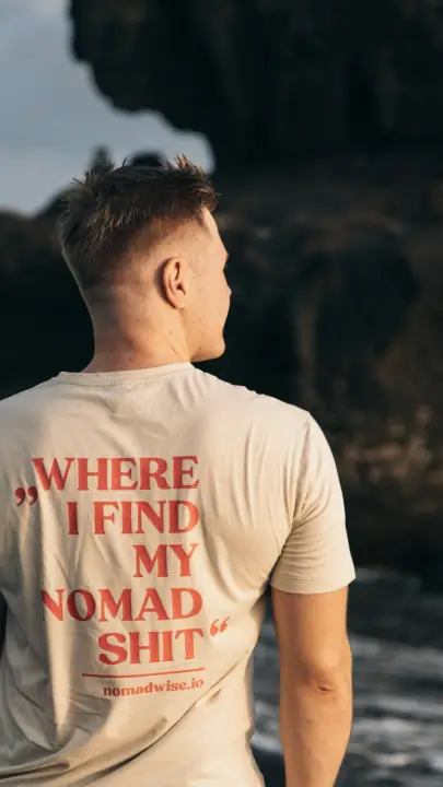 De rugprint Where I find my nomad shit en nomadwise.io in rood op een beige shirt, tegen donkere rotsen