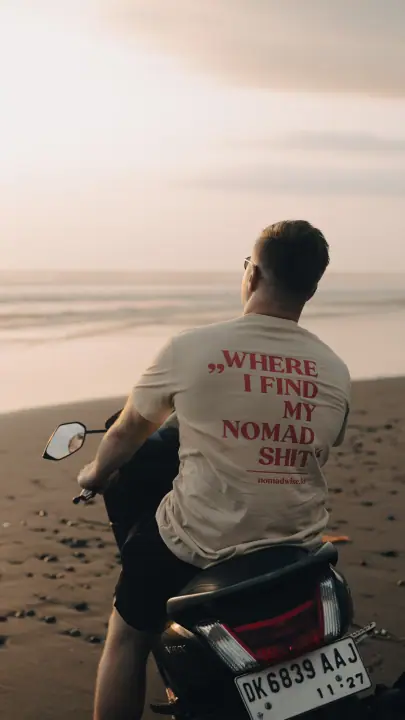 Een man op een scooter kijkt uit over zee bij zonsondergang, de Nomadwise print op zijn beige shirt