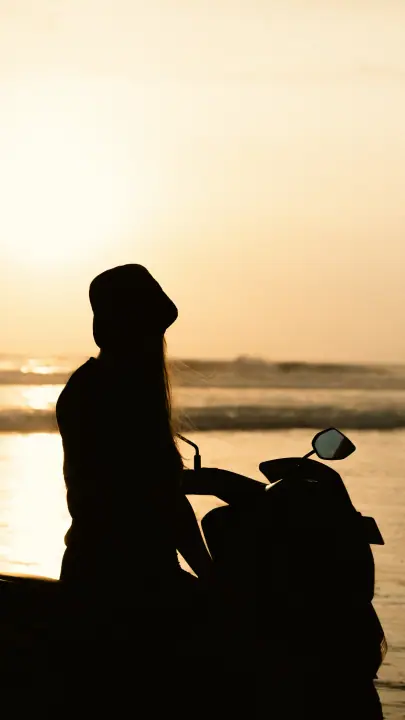 Silhouet van een vrouw met vissershoedje bij haar scooter, de zee goudkleurig in het tegenlicht