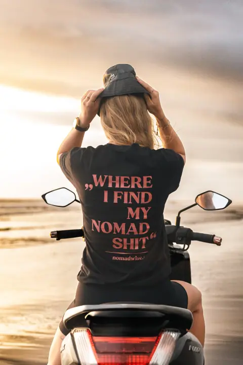Op de scooter aan zee houdt ze haar hoedje vast, Where I find my nomad shit groot op haar rug