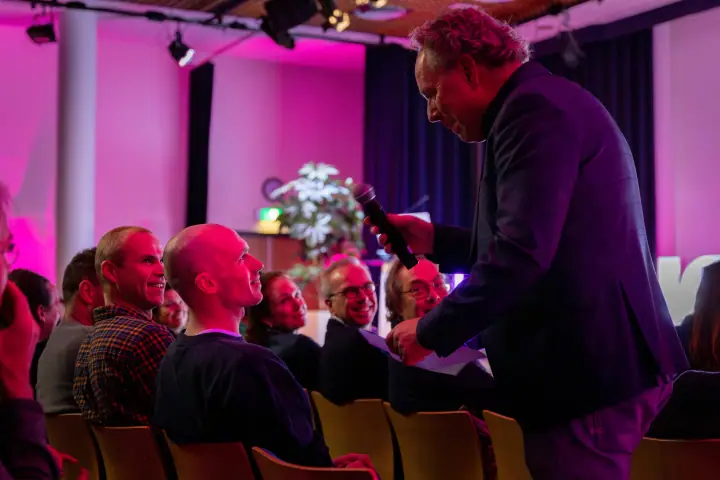 Presentator buigt met de microfoon naar het publiek tijdens een vragenronde, in roze zaallicht