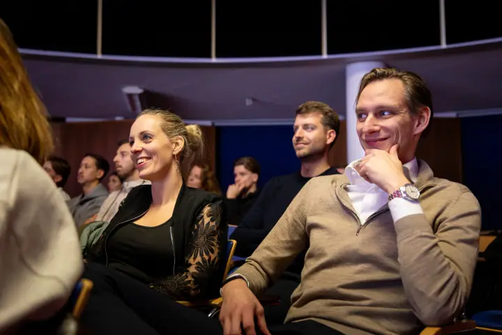 Twee bezoekers glimlachen om een verhaal op het podium, de man met zijn hand onder zijn kin