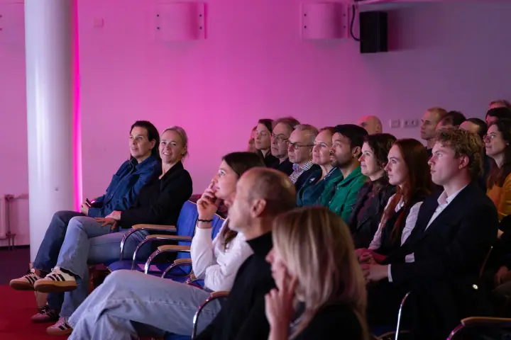 Zijaanzicht van het publiek langs een roze verlichte muur, iedereen gericht op het podium