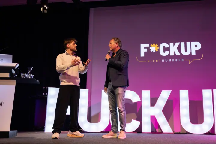 Presentator interviewt een spreker in crèmekleurige trui, met het logo van F*ckup Night Nijmegen erachter